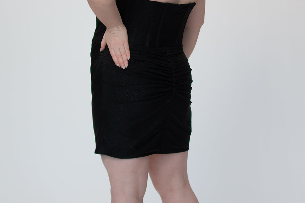 Short fitted black party dress SC8143 by Sydney's Closet. Back view.  Strapless, exposed boning basque waist and ruched fitted skirt in shimmer stretch knit. Availability in black, purple, red, ivory, green and royal. Model in picture is 5'9.