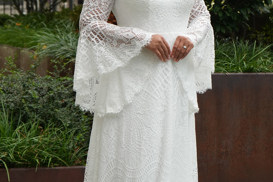 Boho crochet lace plus size wedding dress with V-neckline, illusion lace straps, illusion long flowy bell sleeves, natural waistline and slim A-line skirt finished with scalloped eyelash hem.  Available in ivory. Celebrations by Sydney's Closet style CE2604