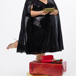Sydney's Closet CE2609 in black.

Plus size sequin black short party dress. V-neckline, bra-friendly straps with attached cape sleeve detail, princess silhouette, slim A-line skirt. at knee-length.

Model posed holding a Christmas present and X-mas gifts on the floor.