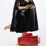 Sydney's Closet CE2609 in black.

Plus size sequin black short party dress. V-neckline, bra-friendly straps with attached cape sleeve detail, princess silhouette, slim A-line skirt. at knee-length.

Model posed holding a Christmas present and X-mas gifts on the floor.