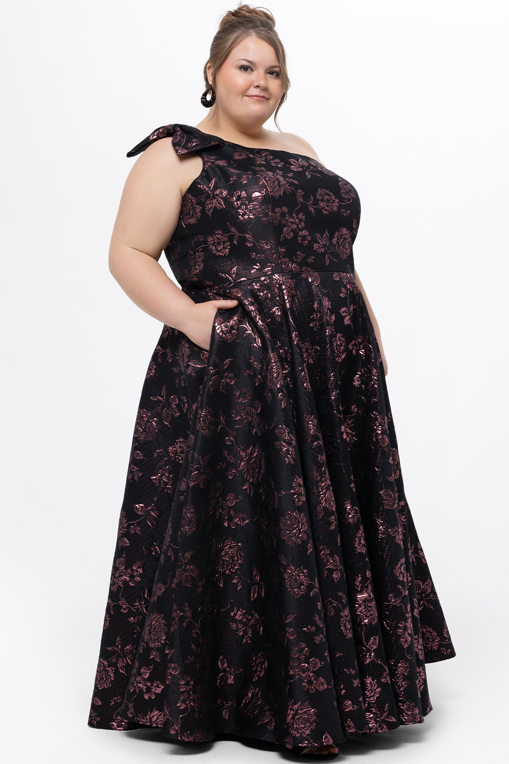 Celebrations by Sydney's Closet Style CE2614 in Pink Rose and Black floral brocade fabric.

Plus size maxi length formal dress. One-shoulder neckline with detachable bow at high point of shoulder, natural waistline and A-line skirt with pockets. 