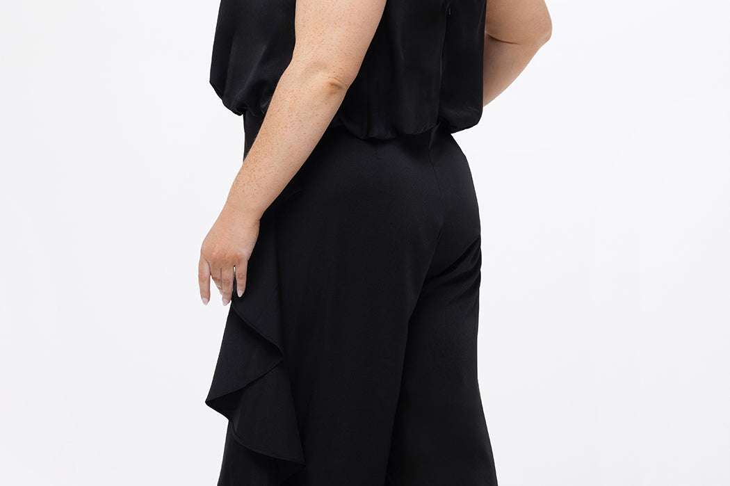 Celebrations by Sydney's Closet Style CE2615 in Black.

One-piece crepe satin jumpsuit with ruffled on outer pants seams. Bra-friendly straps, blouson bodice and wide leg pants. Center back zipper and scoop neckline. 