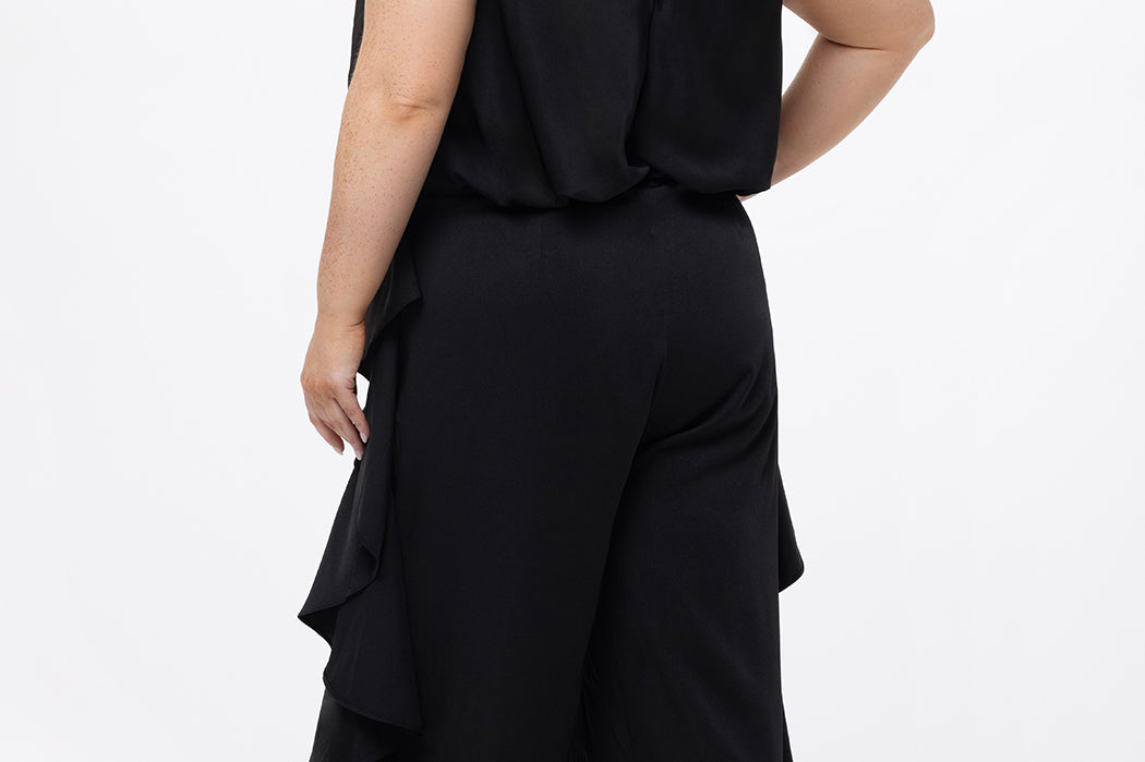 Celebrations by Sydney's Closet Style CE2615 in Black.

One-piece crepe satin jumpsuit with ruffled on outer pants seams. Bra-friendly straps, blouson bodice and wide leg pants. Center back zipper and scoop neckline. 