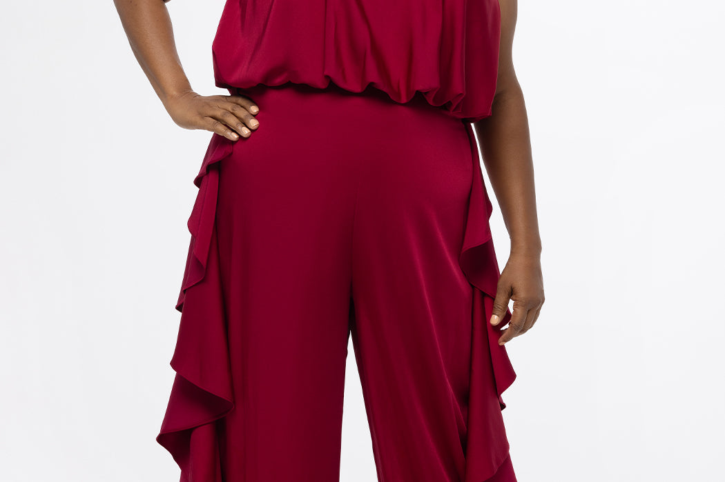 Celebrations by Sydney's Closet Style CE2615 in cherry red.

One-piece crepe satin jumpsuit with ruffled on outer pants seams. Bra-friendly straps, blouson bodice and wide leg pants. Center back zipper and scoop neckline. 