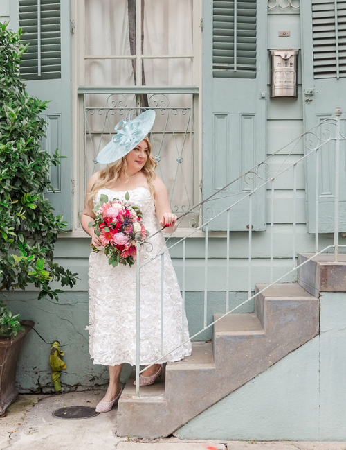 Woman in a white dress with a floral bouquet standing on steps in front of a light blue building.