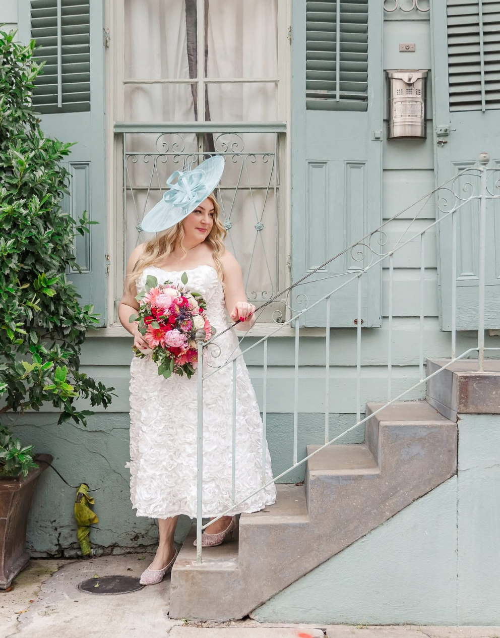Woman in a white dress with a floral bouquet standing on steps in front of a light blue building.