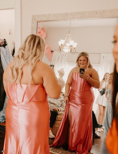 Woman in a coral dress taking a mirror selfie in a room with people admiring in the background.