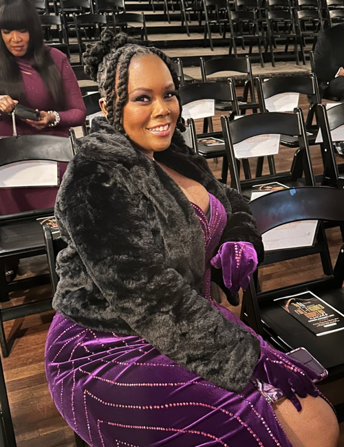 Woman in a purple dress and gloves with a black fur coat sitting in an auditorium.