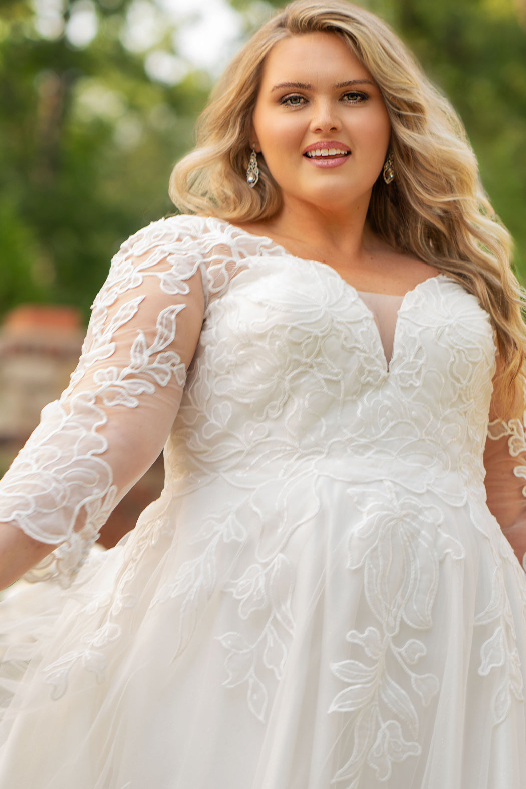 MB2604 by Michelle Bridal

Available in Black, Ivory, Black on Ivory, Ivory on Black, Ivory on Champagne and Black on Champagne. 

Plus-size model wearing an ivory A-line wedding gown with sheer long sleeves, a deep V-neckline, and intricate lace appliqué with subtle shimmer throughout the full skirt, photographed outdoors in front of a stone backdrop.