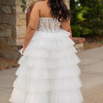 Plus-size bride wearing a strapless white wedding gown with a sheer lace corset bodice and tiered ruffled tulle ball gown skirt, standing outdoors in front of a stone fireplace and greenery.