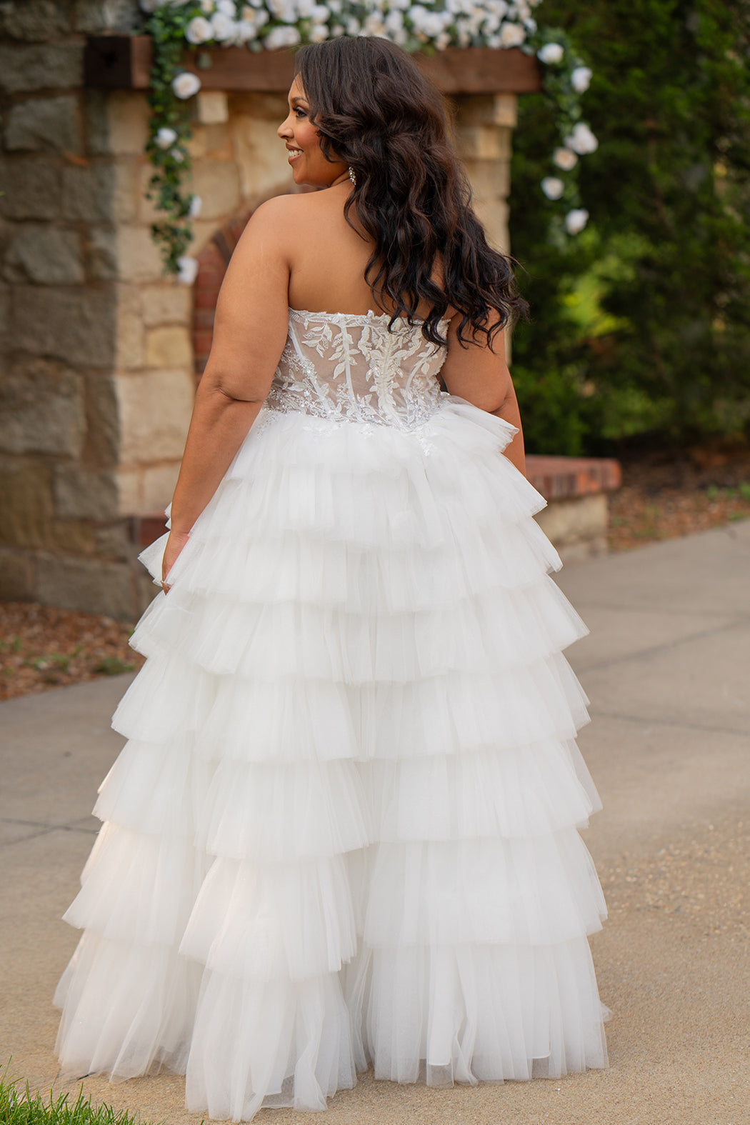 Plus-size bride wearing a strapless white wedding gown with a sheer lace corset bodice and tiered ruffled tulle ball gown skirt, standing outdoors in front of a stone fireplace and greenery.