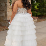 Plus-size bride wearing a strapless white wedding gown with a sheer lace corset bodice and tiered ruffled tulle ball gown skirt, standing outdoors in front of a stone fireplace and greenery.