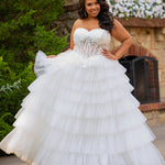 Plus-size bride wearing a strapless white wedding gown with a sheer lace corset bodice and tiered ruffled tulle ball gown skirt, standing outdoors in front of a stone fireplace and greenery.
