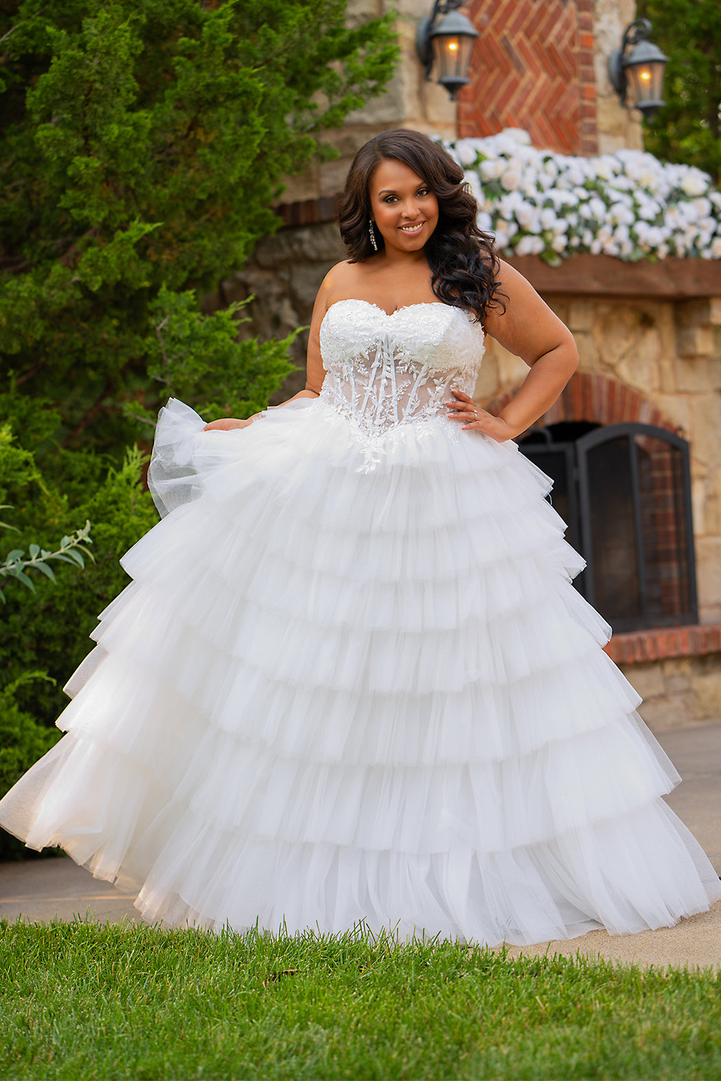 Plus-size bride wearing a strapless white wedding gown with a sheer lace corset bodice and tiered ruffled tulle ball gown skirt, standing outdoors in front of a stone fireplace and greenery.