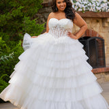 Plus-size bride wearing a strapless white wedding gown with a sheer lace corset bodice and tiered ruffled tulle ball gown skirt, standing outdoors in front of a stone fireplace and greenery.