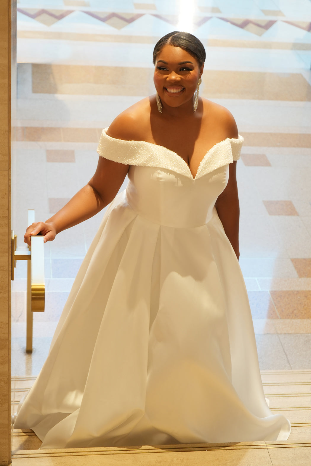 Michelle Bridal MB2610 shown in ivory. Off-the-shoulder satin A-line gown, bodice cuff covered in pearls and clear beads. Natural waistline, box pleats on skirt and center back zipper. Long satin train. 