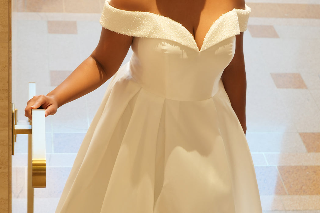 Michelle Bridal MB2610 shown in ivory. Off-the-shoulder satin A-line gown, bodice cuff covered in pearls and clear beads. Natural waistline, box pleats on skirt and center back zipper. Long satin train. 