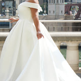 Michelle Bridal MB2610 shown in ivory. Off-the-shoulder satin A-line gown, bodice cuff covered in pearls and clear beads. Natural waistline, box pleats on skirt and center back zipper. Long satin train. 