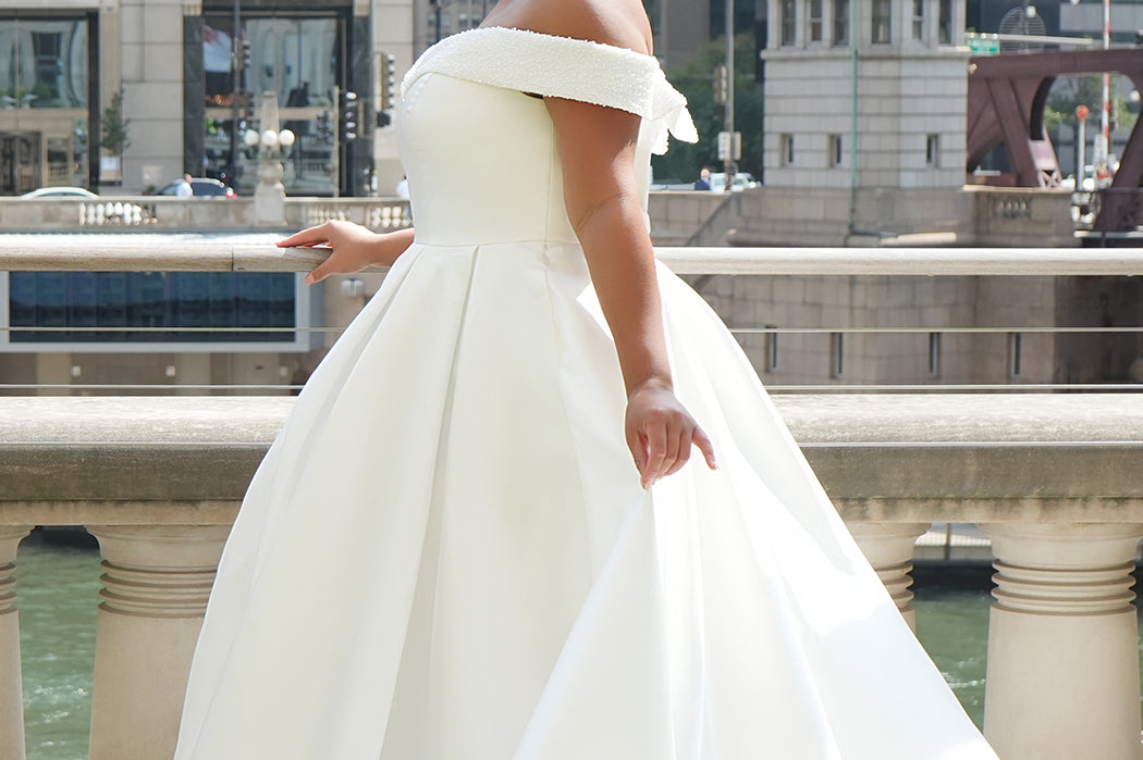 Michelle Bridal MB2610 shown in ivory. Off-the-shoulder satin A-line gown, bodice cuff covered in pearls and clear beads. Natural waistline, box pleats on skirt and center back zipper. Long satin train. 