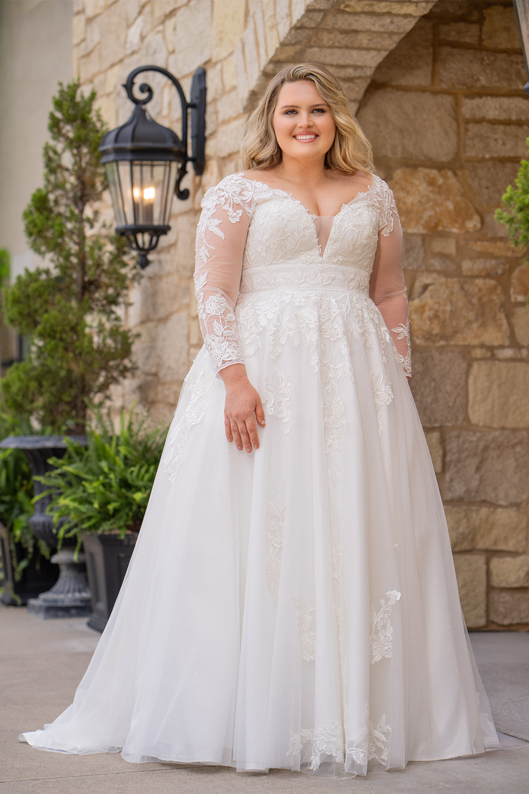 Michelle Bridal MB2615 shown in Ivory. 
Long sleeves covered in large organza appliques, v-neckline, 3 inch waistband. A-line skirt, Soft bridal tulle, large organa appliques, lightweight satin lining. V-Back, Long train and center back zipper. 