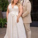 Michelle Bridal MB2625 shown in Ivory. 

Organza brocade A-line silhouette, Pleated faux surplice bodice for elegant shaping, Bra-friendly straps and sleeveless, detachable belt with oversized bow for two looks in one, Box-pleated A-line skirt with front slit for movement, Chapel-length train for timeless finish, Invisible center back zipper.