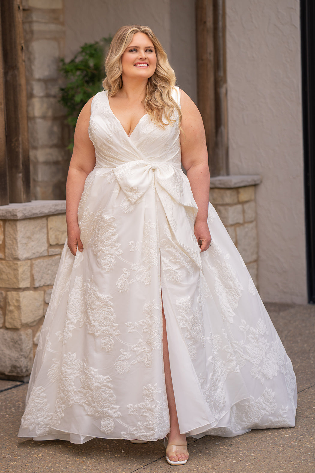 Michelle Bridal MB2625 shown in Ivory. 

Organza brocade A-line silhouette, Pleated faux surplice bodice for elegant shaping, Bra-friendly straps and sleeveless, detachable belt with oversized bow for two looks in one, Box-pleated A-line skirt with front slit for movement, Chapel-length train for timeless finish, Invisible center back zipper.