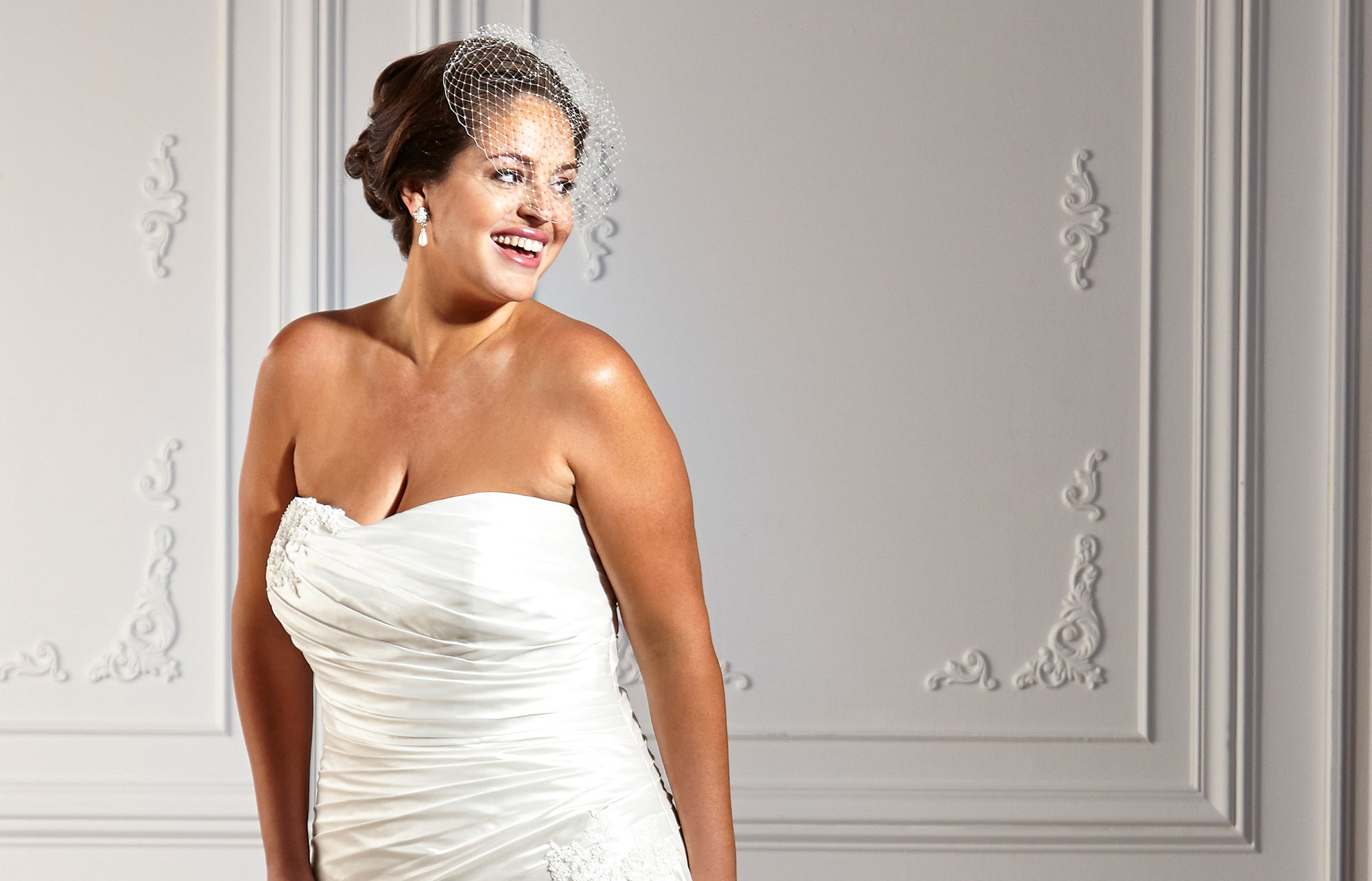 Woman wearing a white strapless wedding dress and a net hair accessory, standing in front of a decorative wall.
