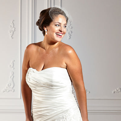 Woman wearing a white strapless wedding dress and a net hair accessory, standing in front of a decorative wall.