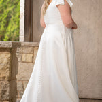 Blonde woman wearing an ivory one-shoulder solid satin wedding down with a bow against a stone wall. 

Sydney's Closet SC5355 ivory plus size A-line wedding dress. One-shoulder neckline, detachable oversized bow on the high point of shoulder and pockets.  Train with buttons down the back. 