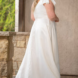 Blonde woman wearing an ivory one-shoulder solid satin wedding down with a bow against a stone wall. 

Sydney's Closet SC5355 ivory plus size A-line wedding dress. One-shoulder neckline, detachable oversized bow on the high point of shoulder and pockets.  Train with buttons down the back. 