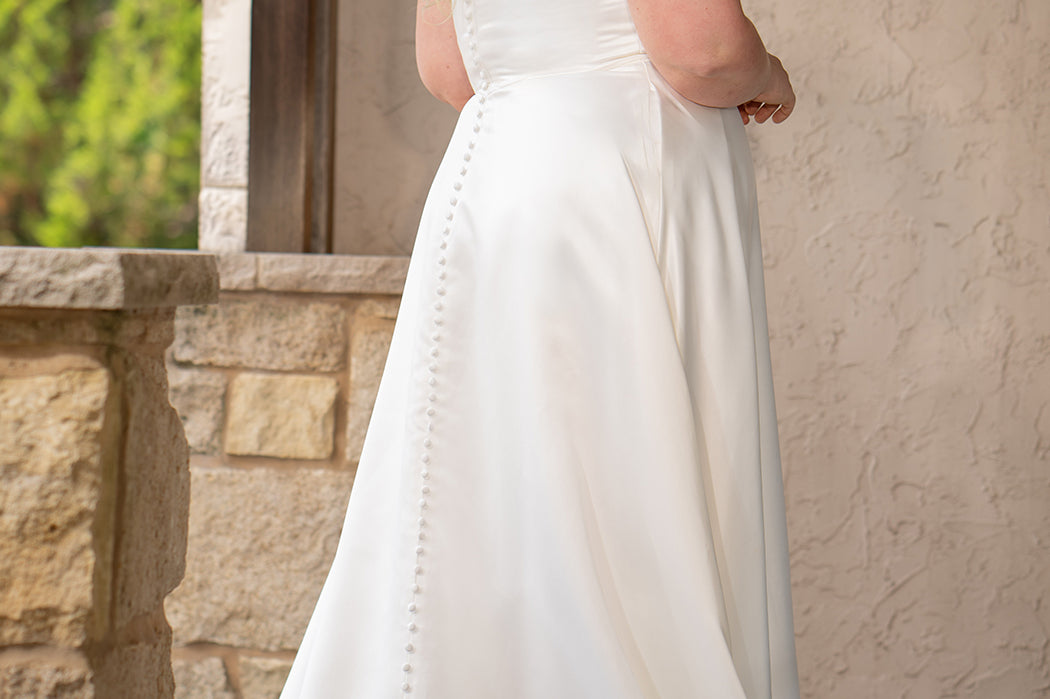 Blonde woman wearing an ivory one-shoulder solid satin wedding down with a bow against a stone wall. 

Sydney's Closet SC5355 ivory plus size A-line wedding dress. One-shoulder neckline, detachable oversized bow on the high point of shoulder and pockets.  Train with buttons down the back. 
