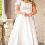Blonde woman wearing an ivory one-shoulder solid satin wedding down with a bow against a stone wall. 

Sydney's Closet SC5355 ivory plus size A-line wedding dress. One-shoulder neckline, detachable oversized bow on the high point of shoulder and pockets.  Train with buttons down the back. 
