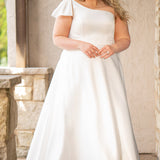 Blonde woman wearing an ivory one-shoulder solid satin wedding down with a bow against a stone wall. 

Sydney's Closet SC5355 ivory plus size A-line wedding dress. One-shoulder neckline, detachable oversized bow on the high point of shoulder and pockets.  Train with buttons down the back. 