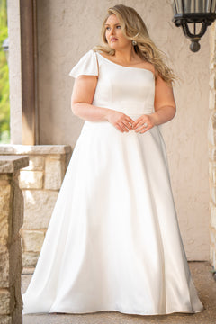 Blonde woman wearing an ivory one-shoulder solid satin wedding down with a bow against a stone wall. 

Sydney's Closet SC5355 ivory plus size A-line wedding dress. One-shoulder neckline, detachable oversized bow on the high point of shoulder and pockets.  Train with buttons down the back. 