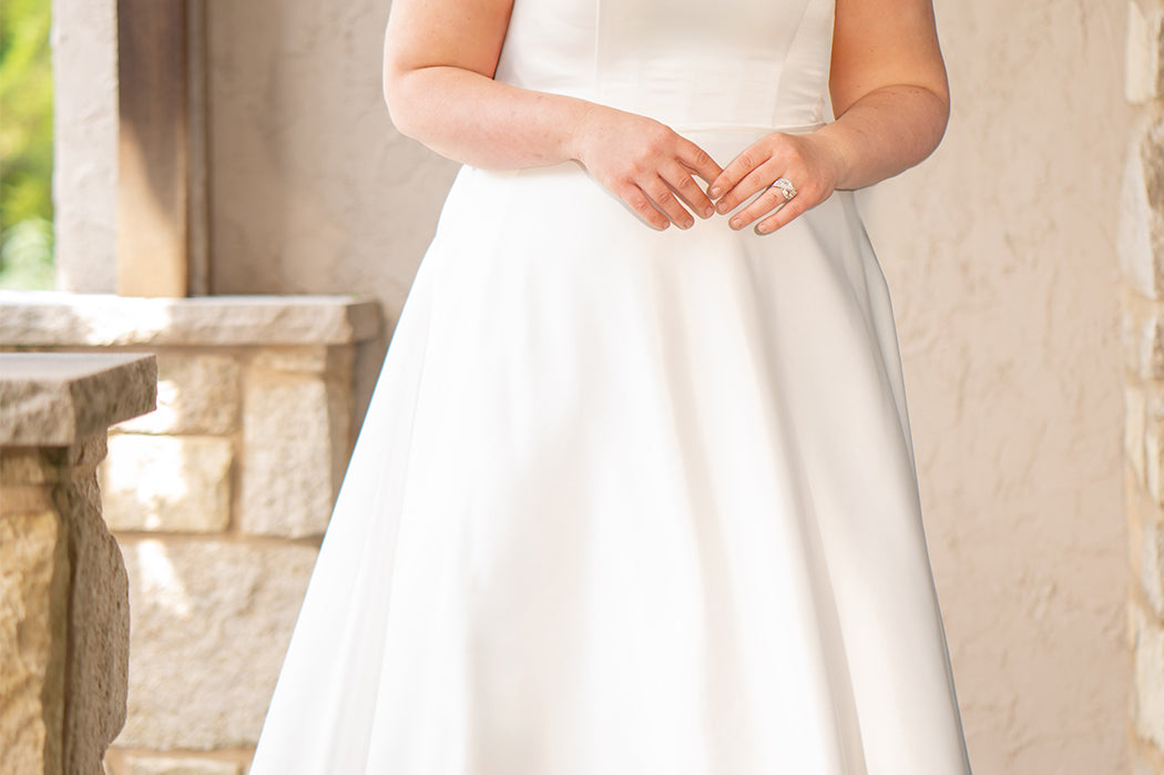 Blonde woman wearing an ivory one-shoulder solid satin wedding down with a bow against a stone wall. 

Sydney's Closet SC5355 ivory plus size A-line wedding dress. One-shoulder neckline, detachable oversized bow on the high point of shoulder and pockets.  Train with buttons down the back. 