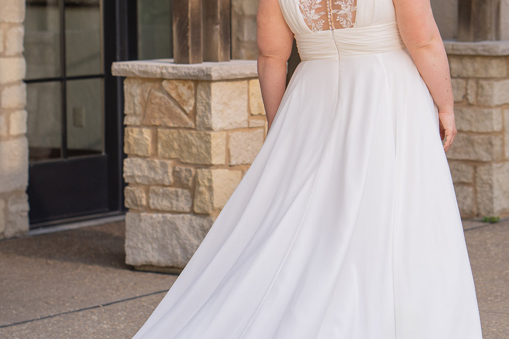 Woman wearing an ivory chiffon wedding dress standing outdoors.

Sydney's Closet style SC5360 in ivory/nude. Plus size light flowy chiffon bridal dress with pleated bodice and pleated waistband.  Illusion back covered in lace appliques with crystal buttons. Available in sizes 14-40. 