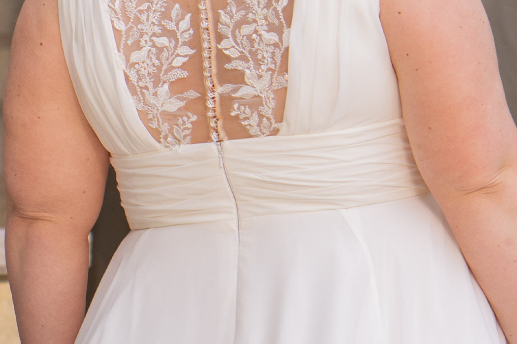 Woman wearing an ivory chiffon wedding dress standing outdoors.

Sydney's Closet style SC5360 in ivory/nude. Plus size light flowy chiffon bridal dress with pleated bodice and pleated waistband.  Illusion back covered in lace appliques with crystal buttons. Available in sizes 14-40. 