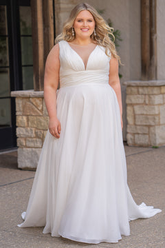 Woman wearing an ivory chiffon wedding dress standing outdoors.

Sydney's Closet style SC5360 in ivory/nude. Plus size light flowy chiffon bridal dress with pleated bodice and pleated waistband.  Illusion back covered in lace appliques with crystal buttons. Available in sizes 14-40. 