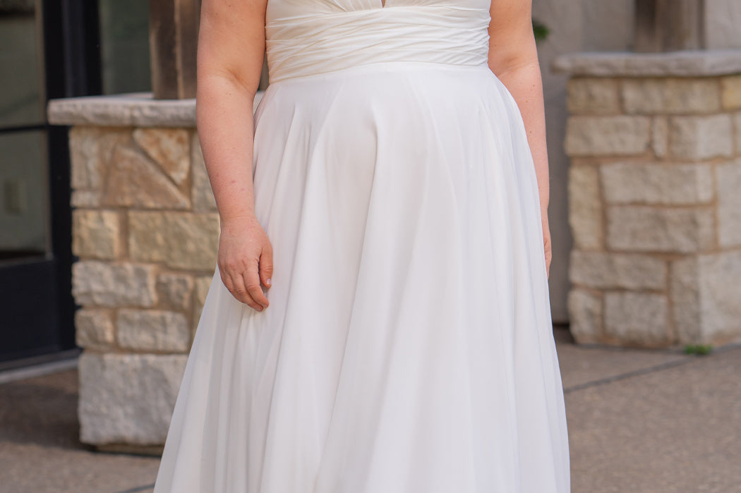 Woman wearing an ivory chiffon wedding dress standing outdoors.

Sydney's Closet style SC5360 in ivory/nude. Plus size light flowy chiffon bridal dress with pleated bodice and pleated waistband.  Illusion back covered in lace appliques with crystal buttons. Available in sizes 14-40. 