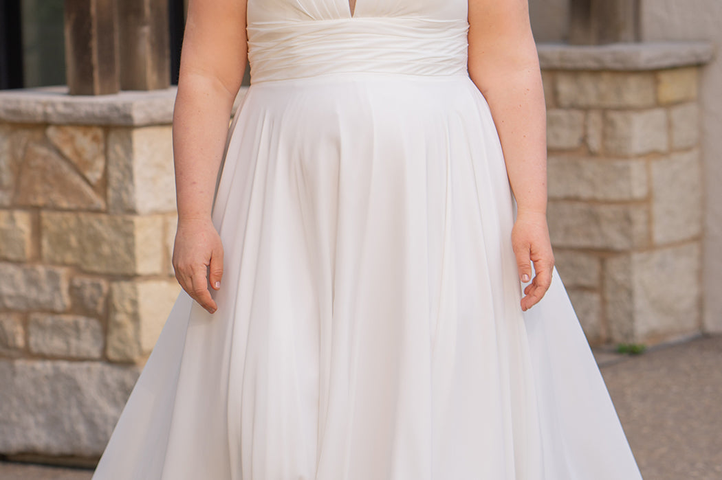 Woman wearing an ivory chiffon wedding dress standing outdoors.

Sydney's Closet style SC5360 in ivory/nude. Plus size light flowy chiffon bridal dress with pleated bodice and pleated waistband.  Illusion back covered in lace appliques with crystal buttons. Available in sizes 14-40. 
