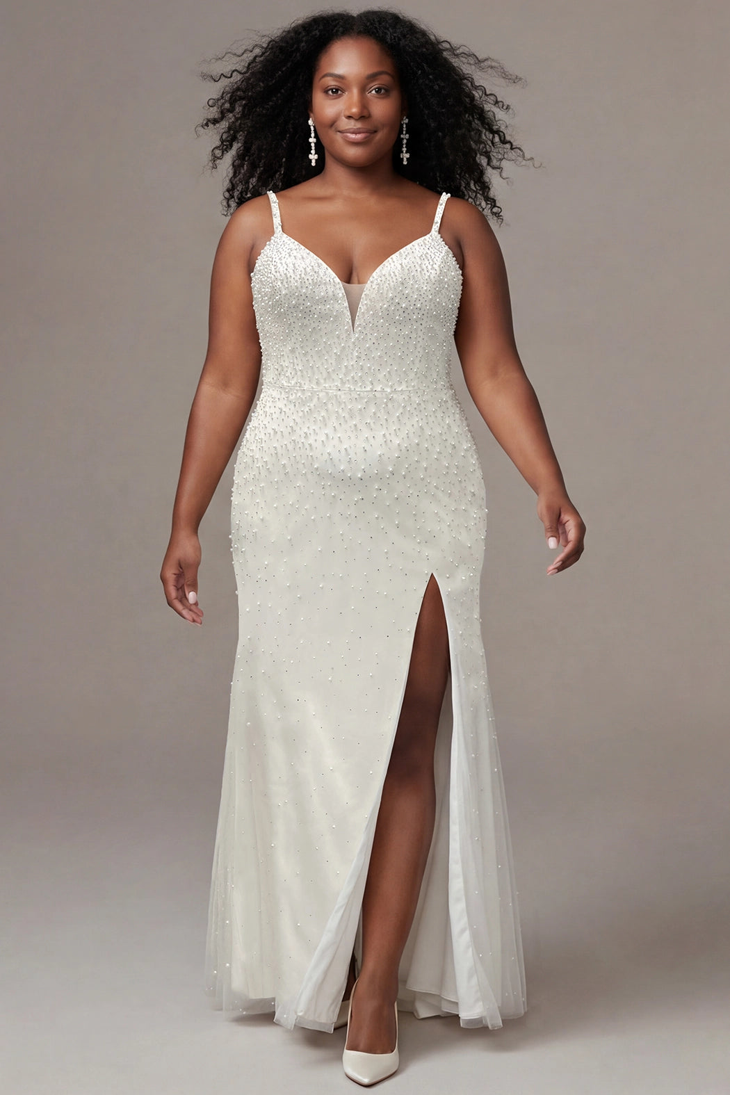 Plus-size bride wearing an elegant ivory wedding gown with a fitted silhouette, delicate spaghetti straps, and a plunging V-neckline with sheer nude inset detail. The bridal dress features allover pearls and silver stone embellishments, a defined waist, and a floor-length skirt with a thigh-high slit. She is styled with natural curls, sparkling drop earrings, and ivory pointed-toe heels, walking confidently against a neutral studio background.

Sydney's Closet SC5377