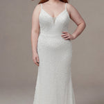 Plus-size bride wearing a fitted ivory lace wedding gown with delicate spaghetti straps, a plunging V-neckline with a sheer inset, and allover pearl-inspired beadwork. The figure-flattering silhouette contours the body with appliques on the side seams before gently flaring into a soft sweep train, creating an elegant and timeless bridal look. She is styled with long red hair and sparkling drop earrings, posing against a neutral studio background.

Sydney's Closet SC5382