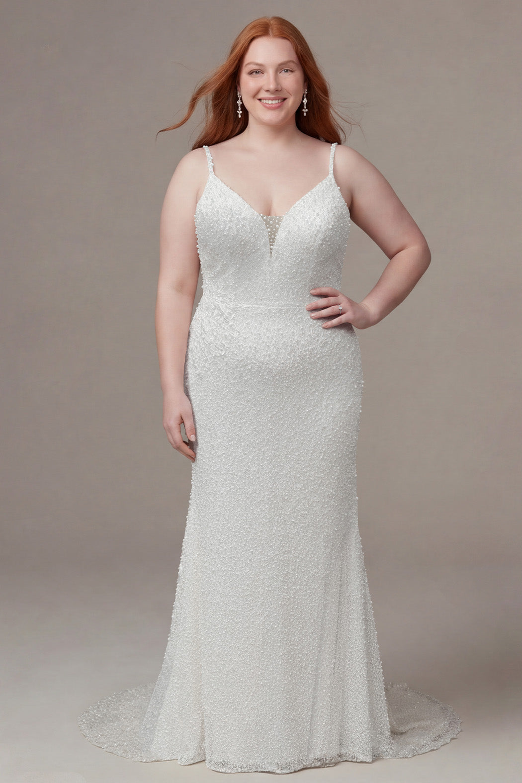 Plus-size bride wearing a fitted ivory lace wedding gown with delicate spaghetti straps, a plunging V-neckline with a sheer inset, and allover pearl-inspired beadwork. The figure-flattering silhouette contours the body with appliques on the side seams before gently flaring into a soft sweep train, creating an elegant and timeless bridal look. She is styled with long red hair and sparkling drop earrings, posing against a neutral studio background.

Sydney's Closet SC5382