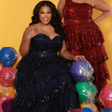 SC7398 & SC7399 duo picture with oversized candy for prom 2025. 

Strapless, sweetheart plus size gala formal ballgown. Exposed corset boning on bodice covered in lace applqiues. Tiered A-line skirt with matching lace applique hem. Style SC7398 is available in forest green, navy blue or burgundy red by Sydney's Closet. 

Plus size one-shoulder formal gala Mardi Gras gown with embroidered sequin lace appliques on bodice and on tiered A-line skirt with slit. Available in forest green. navy blue and burgundy r