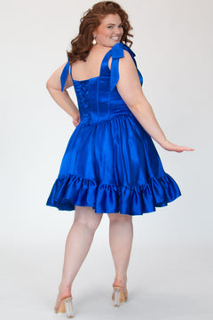 SC8152 by Sydney's Closet. Sapphire blue, back view. A short satin party dress with a 5 inch ruffle at the hem and decorative bows at the top of the shoulder. A lace up back, exposed boning and sweetheart neckline. With a basque waist and lace up back. Available in Sapphire blue and Lipstick pink, sizes 14-28. 
Model is 5'9. 