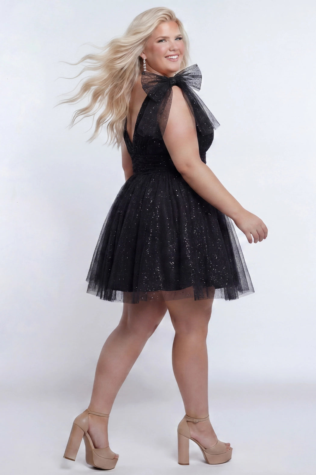 Short A-line plus size flowy party dress SC8153 by Sydney's Closet in the color Onyx black. Back view. Layers of dense sparkle tulle and a satin lining. V-neckline with thick bra-friendly straps and detachable bows at the top of the shoulder. Pleated bodice and 3 inch pleated waistband. Pockets and long invisible center back zipper. Available in Shamrock green, Admiral blue, Firecracker red and Shamrock green in sizes 14-32. Model is 5'9. 
