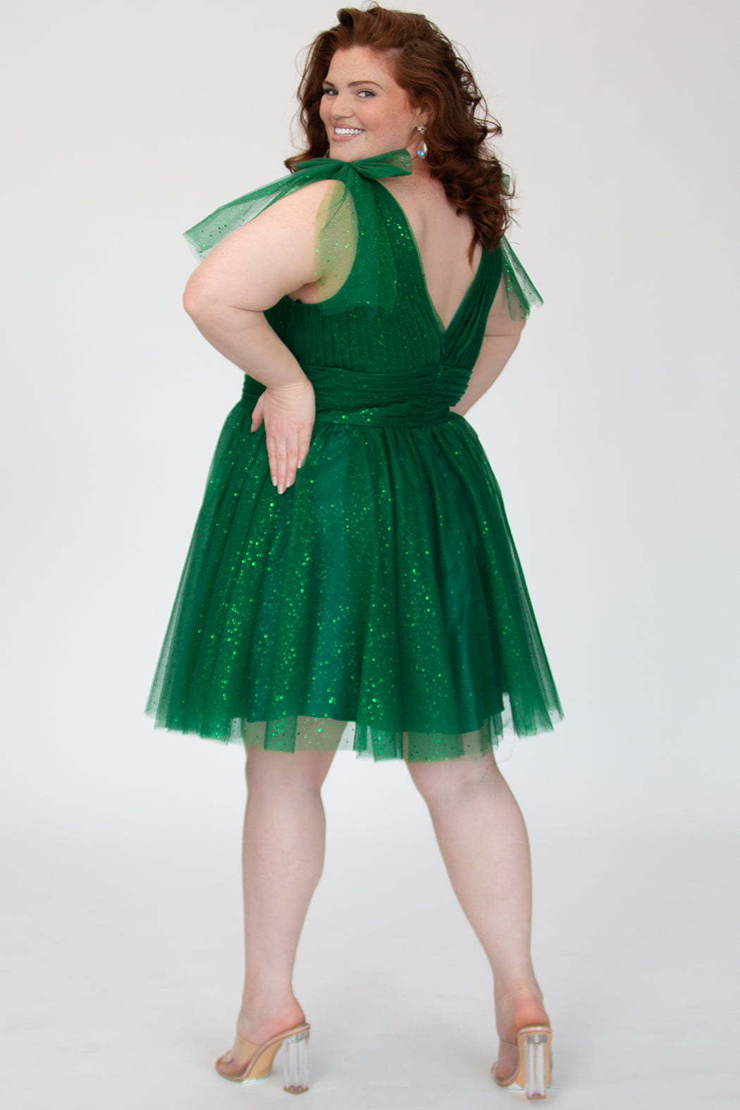 Short A-line plus size flowy party dress SC8153 by Sydney's Closet in the color Shamrock green. Back view. Layers of dense sparkle tulle and a satin lining. V-neckline with thick bra-friendly straps and detachable bows at the top of the shoulder. Pleated bodice and 3 inch pleated waistband. Pockets and long invisible center back zipper. Available in Shamrock green, Admiral blue, Firecracker red and Onyx black in sizes 14-32. Model is 5'9. 
