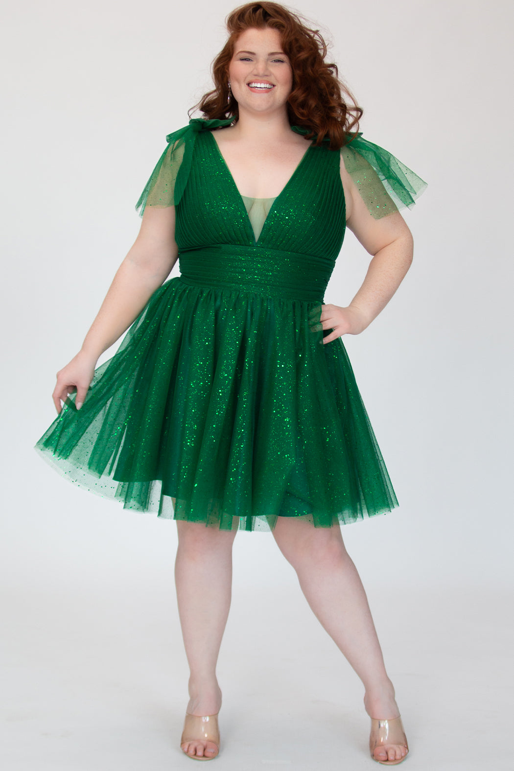 Short A-line plus size flowy party dress SC8153 by Sydney's Closet in the color Shamrock green. Front view. Layers of dense sparkle tulle and a satin lining. V-neckline with thick bra-friendly straps and detachable bows at the top of the shoulder. Pleated bodice and 3 inch pleated waistband. Pockets and long invisible center back zipper. Available in Shamrock green, Admiral blue, Firecracker red and Onyx black in sizes 14-32. Model is 5'9. 