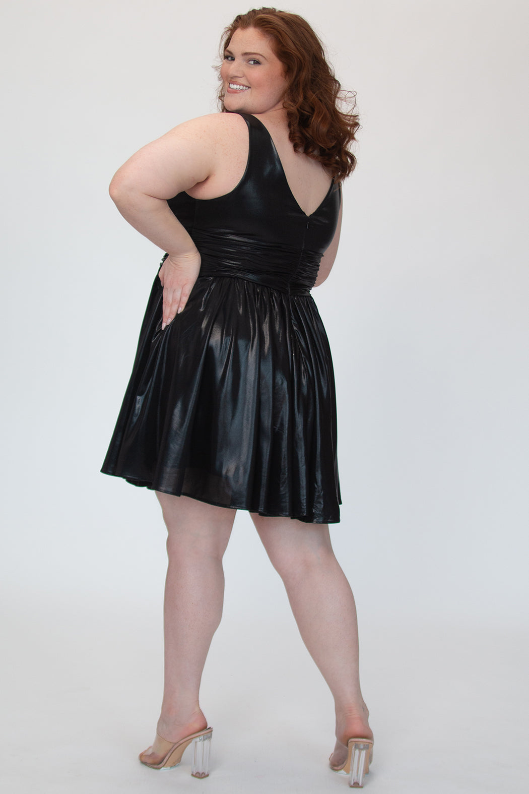 A-line flowy short party dress, SC8156 by Sydney's Closet. Back view. In the color raven black, a  metallic knit fabric. Thick bra-friendly straps, pleated v-neckline bodice and waist band, long invisible center back zipper. V- back. Available in Prism, Peacock and Raven black, sizes 14-32. 
Model in picture is 5'9. 
