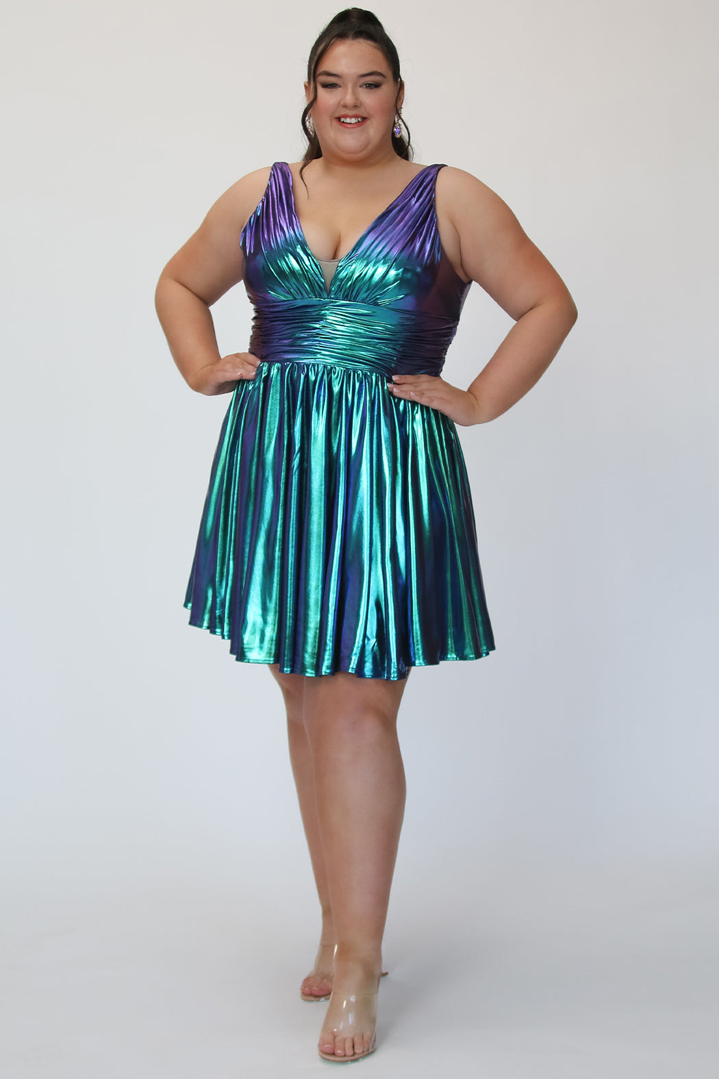 A-line flowy short party dress, SC8156 by Sydney's Closet. Front view. In the color Peacock, a  blue/green metallic knit fabric. Thick bra-friendly straps, pleated v-neckline bodice and waist band, long invisible center back zipper. V- back. Available in Prism, Peacock and Raven black, sizes 14-32. 
Model in picture is 5'9. 